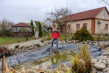 Beyaz, orta yaşlı bir adam bir bahçe havuzunu balçık, su bitkileri, düşen yapraklar ve balık ağlarıyla temizliyor.