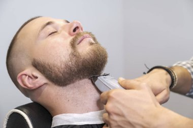 Bir berber müşterisinin sakalını elektrikli makasla şekillendirir. Müşteri gözleri kapalı, seçici bir şekilde oturuyor.