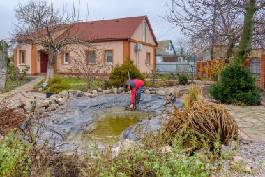 Bir adam bahçe havuzunu balçıktan, su bitkilerinden, düşen yapraklardan bir ağ ile temizler ve balık yakalar.