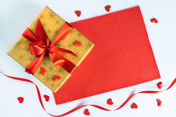 Goldene Geschenkschachtel mit roter Schleife auf Stoff und kleinen Herzen, Konzept des Valentinstages — Stockfoto