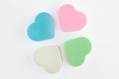 Beyaz arkaplanda kalp şeklinde çeşitli pembe, mavi, yeşil ve beyaz pastel kozmetik süngerler