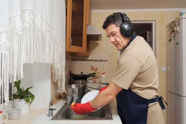 Кавказский мужчина средних лет в красных резиновых перчатках моет посуду губкой на кухне и слушает музыку — стоковое фото