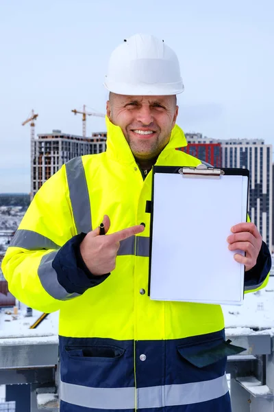 Budowniczy w białym, twardym kapeluszu i żółtej kamizelce trzyma schowek i wskazuje palcem na pustą kartkę papieru — Zdjęcie stockowe