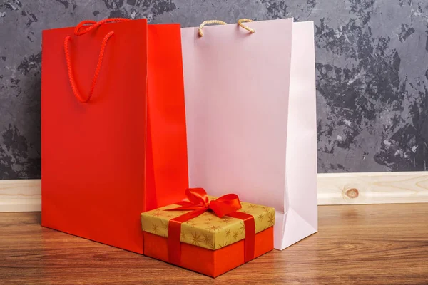 Rote und rosa Papiereinkaufstaschen und goldene Geschenkschachteln auf dem Holzboden gegen eine Betonwand — Stockfoto