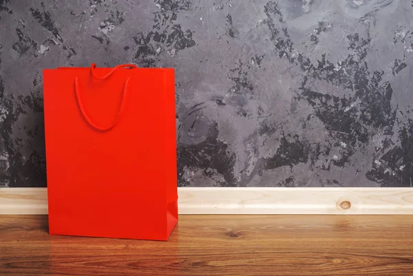 Piros papír bevásárló táska a fa padló ellen beton fal háttér. — Stock Fotó