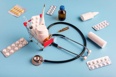 İlaç, şırınga ve termometre ile alışveriş arabası. Steteskop, çeşitli ilaçlar, sprey, mavi arka planda mini sepetin yanına saçılmış haplar. Eczane Alışveriş Konsepti