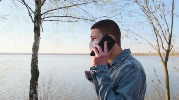 Niño en chaqueta vaqueros lleva máscara protectora, hablando de teléfono móvil y caminando en el lago frente al mar en el parque, vista lateral . — Vídeos de Stock