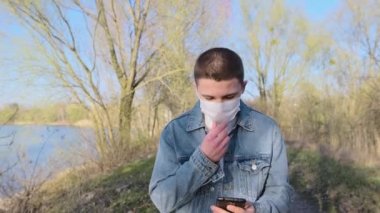 Kot ceketli genç koruyucu maske takıyor, cep telefonuyla konuşuyor ve parktaki göl kenarında yürüyor. İlkbahar, yakın çekim, yavaş çekim.