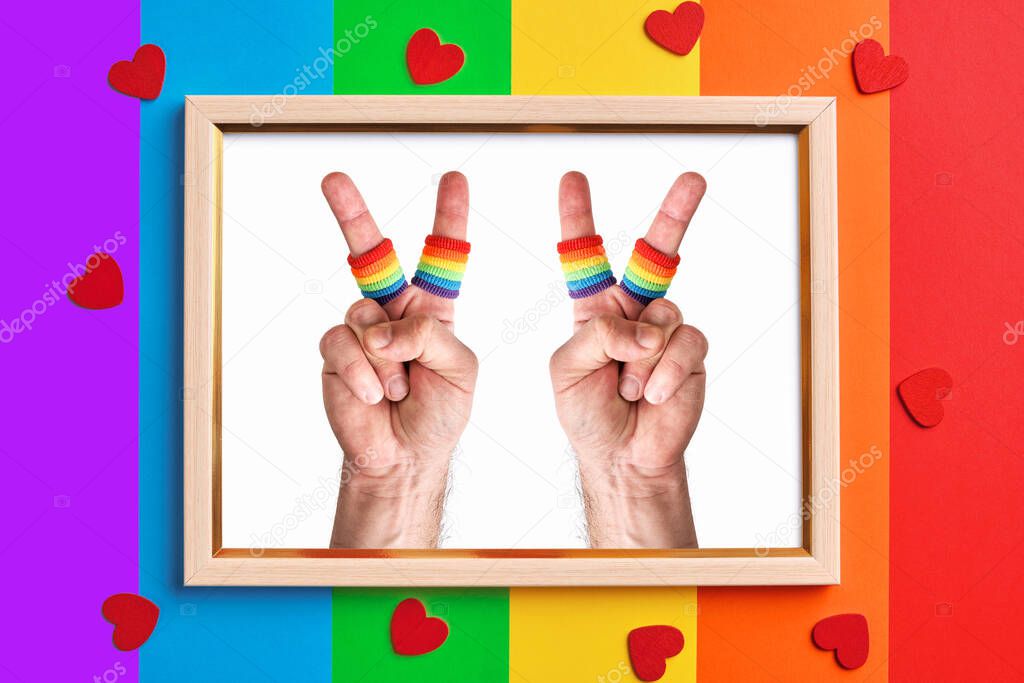 wooden frame with two male hands making the V sign on multicolored rainbow flag background and little red hearts. fingers wears wristbands painted as rainbow flag. Pride day concept, flat lay