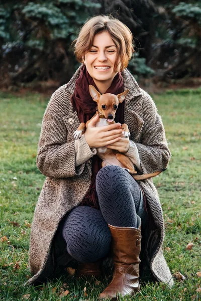 Sorridente ragazza in cappotto tiene un piccolo cane giocattolo Terrier tra le braccia in autunno. cane mostra lingua . — Foto Stock