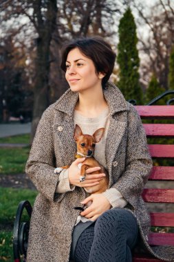 brunette young woman holds small toy Terrier dog in her arms while sitting on red bench in autumn Park