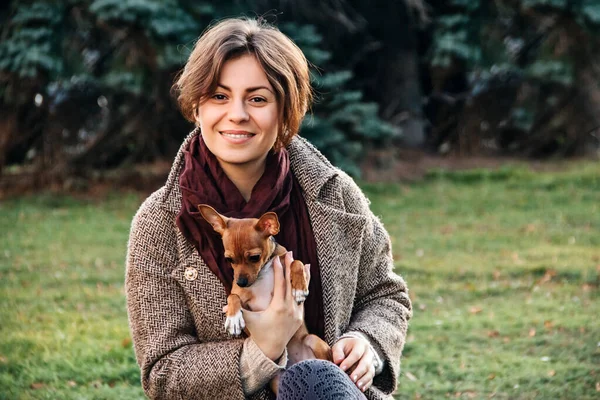 Giovane donna in cappotto tiene un piccolo cane giocattolo Terrier tra le braccia in autunno . — Foto Stock