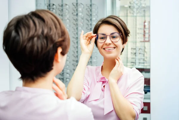 Femeie tânără în bluză roz încercând ochelari la modă în magazinul optometrist uitându-se la ea însăși în oglindă — Fotografie, imagine de stoc