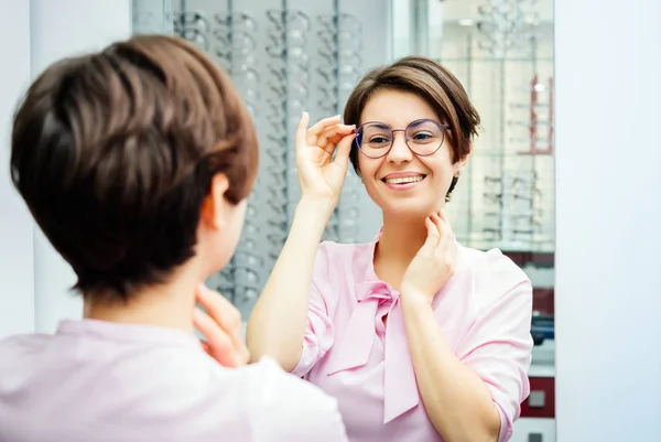 Pembe bluzlu genç bir kadın optometrist dükkanında şık gözlükler deniyor. Aynada kendine bakıyor.