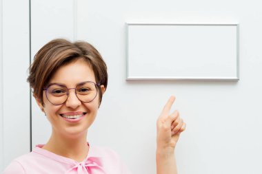 Genç bir esmer kadın, gözlükleriyle gülümsüyor ve parmağını beyaz kapıdaki tabelaya doğrultuyor.