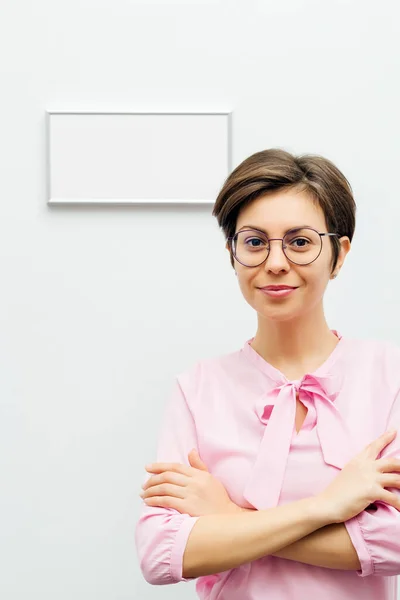 An attractive smiling young brunette stands with her arms folded against a white wall with a sign on it Stock Image