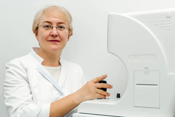 Una mujer adulta con una bata blanca detrás de un dispositivo oftalmológico mira a la cámara — Foto de Stock