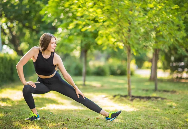 Büyük yeşil parkta uzanan büyüleyici uzun hafif saçlı gülümseyen bir kadın yapıyor. Sportif Bayan siyah topaç, siyah tayt ve yeşil desenli siyah antrenman ör içinde giyinmiş.