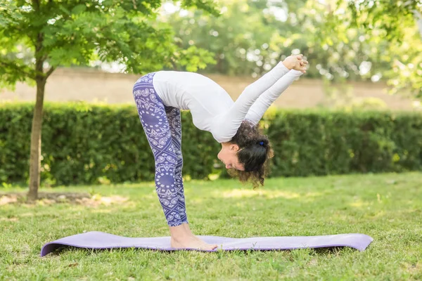 The wonderful and cute woman practicing yoga pose in the park with a lot of trees. A charming lady is doing sports exercises in a white sweater and blue leggings.