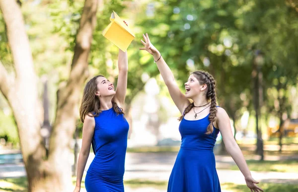 Twee fantastische en mooie meisjes in licht blauw jurken zijn gelukkig en glimlachend in het grote park. Het schattige meisje met een gele boek in haar hand. — Stockfoto