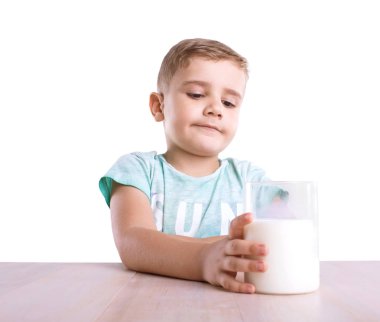 Kendinden emin ve yakışıklı küçük bir çocuk sarı saçlı ve mavi T-shirt tutan bir bardak ile parlak bir ahşap masa, lezzetli süt dolu bir izole beyaz bir arka plan üzerinde. 