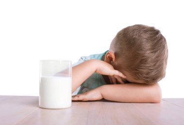 Küçük şirin üzgün bir çocuk mavi tişört, beyaz bir arka plan üzerinde izole ahşap masa lezzetli süt dolu cam yakın oturuyor. 