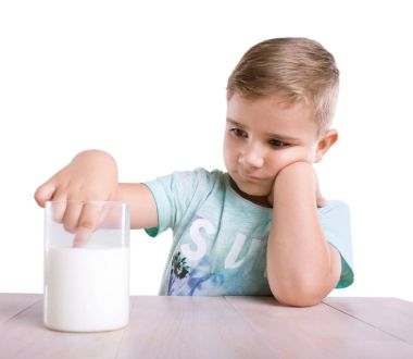 Yakışıklı bir oğlan mavi tişört, beyaz bir arka plan üzerinde izole ahşap masa üzerinde organik süt dolu büyük cam içine onun parmağı dips.