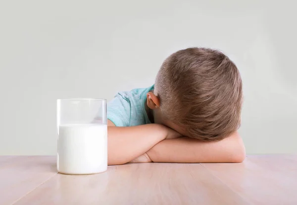 A glass of white nutritious milk next to the sad adorable little kid on the light brown wooden table. Lactose intolerance. — Stock Photo, Image