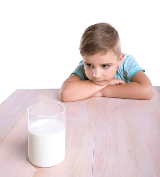 Egy vicces aranyos kisfiú a kék pólóban fekszik, egy nagy, világos fa asztal és néz egy hatalmas üveg tele finom tejet, elszigetelt fehér background. — Stock Fotó