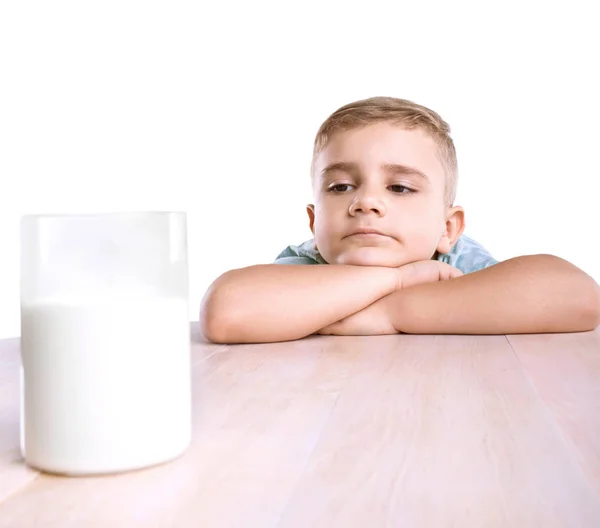 Un niño lindo en una camiseta azul se encuentra con un enorme vaso de leche orgánica. Un niño lindo mira el vaso de leche de buen gusto en una mesa de madera marrón, aislado sobre un fondo blanco . —  Fotos de Stock