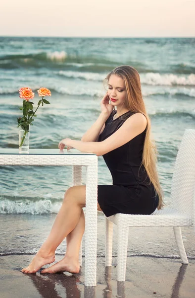 Volledige lengte van een charmant blond meisje in een modieuze en zwarte jurk op de achtergrond van de zee. Een leuke jonge dame op de achtergrond van de zee. — Stockfoto