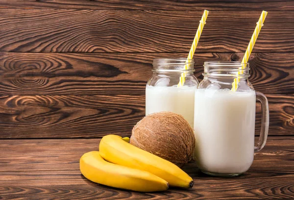 Trópusi gyümölcsöket és üveg tejet a barna háttér. Mason tégelyek, teljes tej, a sorsot. Egzotikus kókusz. Egészséges banán. — Stock Fotó