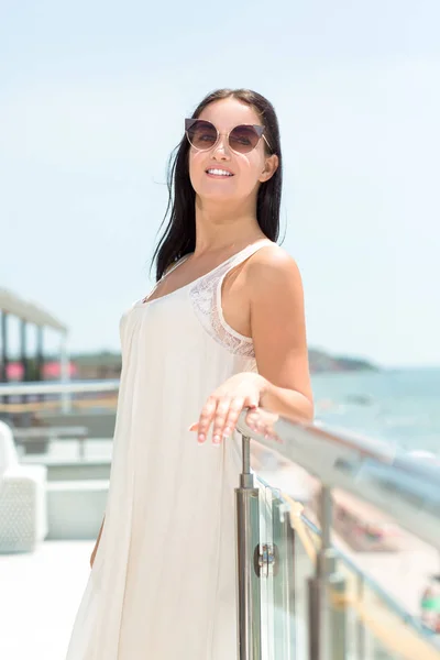 Una donna elegante in un vestito su uno sfondo cielo. Una signora attraente in piedi su un balcone dell'hotel. Una ragazza che guarda l'acqua fresca dell'oceano. Una femmina felice con gli occhiali da sole . — Foto Stock