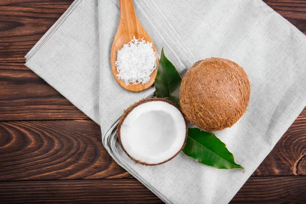 Coco blanco de buen gusto agrietado por la mitad y un delicioso coco entero con hojas verdes y una cuchara llena de chips de nuez fresca en una mesa de madera. Frutas tropicales . — Foto de Stock