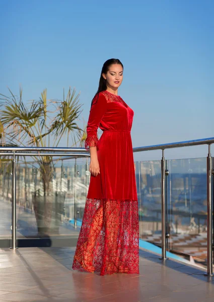 Uma senhora morena de pé em uma varanda em um fundo céu azul. Uma mulher na moda em um vestido vermelho longo. Uma fêmea de férias . — Fotografia de Stock