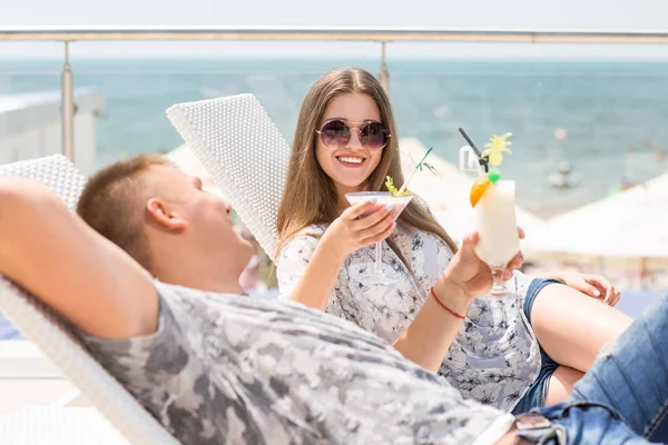 Le giovani coppie si rilassano nella zona estiva dell'hotel. Affascinante, dolce e amorevole coppia sdraiata su sedie a sdraio, bere bevande colorate e rinfrescanti e sorridere carino tra di loro . — Foto Stock