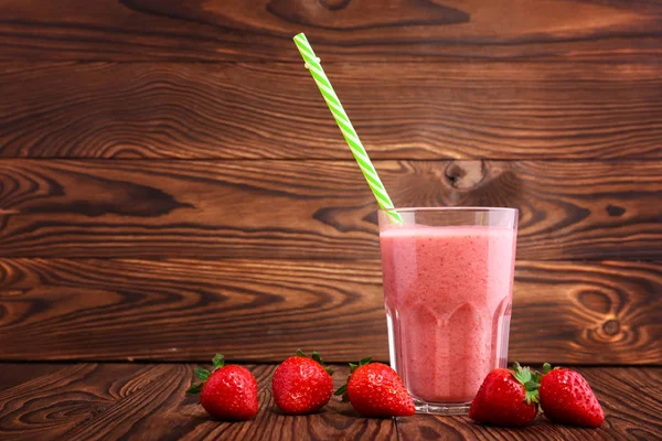 Ein Glas frischen und süßen Smoothie auf einem braunen Holztisch. ein dünner und gerade grüner Strohhalm im transparenten Glas mit dem Getränk. ein paar Erdbeeren sind in der Nähe eines vollen Glases Getränk. — Stockfoto