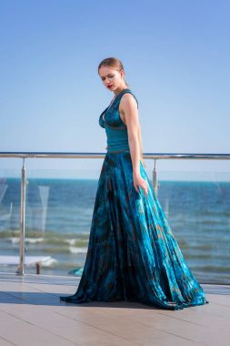 Çekici ve genç bir kadın bir cam balkonda kalıyor. Parlak mavi deniz arka plan üzerinde turkuaz elbiseli çekici bir kız.