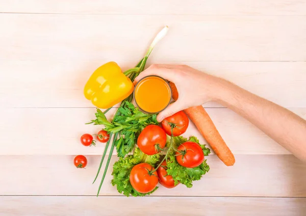 Różnych warzywa: papryka, pomidory, marchew, sałata i pietruszka. Sałatki na jasny drewniany stół. Ręka z szklankę soku. — Zdjęcie stockowe