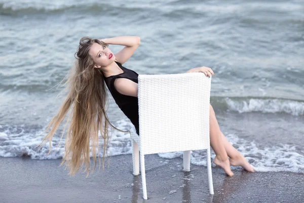 En fantastisk tjej på naturliga blå havet bakgrund. En hot damen i en smal svart klänning. En snygg kvinna som sitter på en stol. — Stockfoto