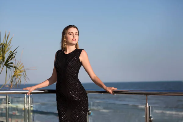 Una donna lussuosa su uno sfondo blu cielo. Una ragazza con un vestito nero magro in piedi su un balcone. Una splendida vista sull'oceano . — Foto Stock