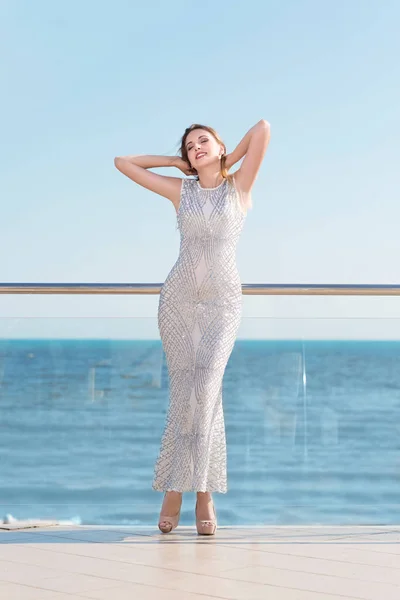 Een aantrekkelijke vrouw in een fantastische witte jurk op een achtergrond van de heldere blauwe lawaaierige zee. De jonge vrouw op een zomervakantie. Het prachtige meisje poseren op een terras. — Stockfoto