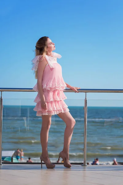 Egy vonzó és gyönyörű nő egy rózsaszín ruhában pózol, egy napos szálloda erkélyén. A csodálatos lány egy világos rózsaszín ruha ruches a tenger közelében. A fényűző életmód. — Stock Fotó