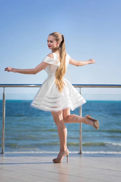 Μια όμορφη κυρία, το περπάτημα σε μια βεράντα. Ένα χαμογελαστό γυναίκα με μακριά μαλλιά σε φωτεινό ουρανό φόντο. Ένα κομψό κορίτσι σε ένα κοντό λευκό φόρεμα κοντά στη θάλασσα. — Φωτογραφία Αρχείου