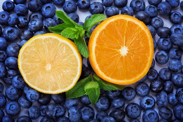 Top view of fresh and juicy halves of lemon and orange on a blueberry background. Summer fruits. Ripe blueberry as texture background and a bright yellow halves of lemon and orange with mint. — Stock Photo, Image