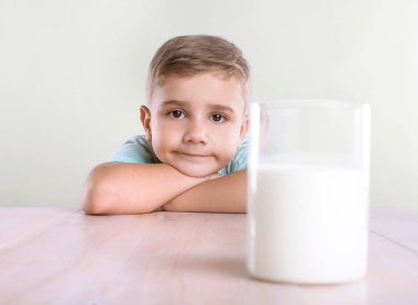 Güzel bir çocuk ona bir bardak süt büyük bir ahşap kahverengi tabloda yakınlarında ve gülümsüyor. Bir bardak süt, beyaz bir arka plan üzerinde izole olan mavi bir tişört meraklı küçük çocukta. 