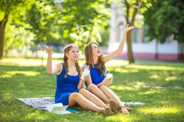 Twee waanzinnig mooie meisjes zijn zit in de zomer in het park en het gooien van laptops. Mooie jonge meisjes in blauwe jurken buiten. — Stockfoto