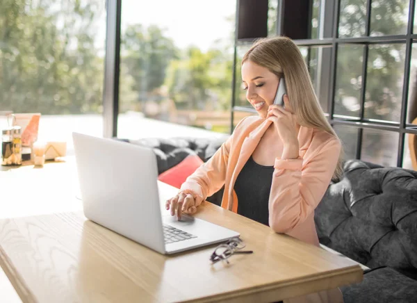 Office giysili bir genç ve çekici iş kadını masada oturan ve telefonda konuşurken ve laptop arıyorum. Başarılı ve güzel genç iş kadını ofiste. — Stok fotoğraf