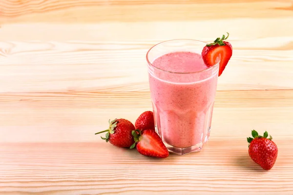 Ett stort glas fyllt med en smakfull milkshake med en röd jordgubbe på toppen på en ljus brun trä skrivbord bredvid några söta jordgubbar. — Stockfoto