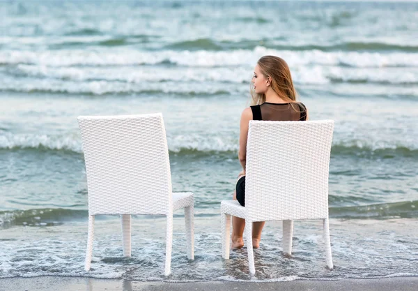 Egy gyönyörű lány ül egy fehér széket, és a tengerre néz. A bájos fiatal lány ül a háttérben a tenger. — Stock Fotó